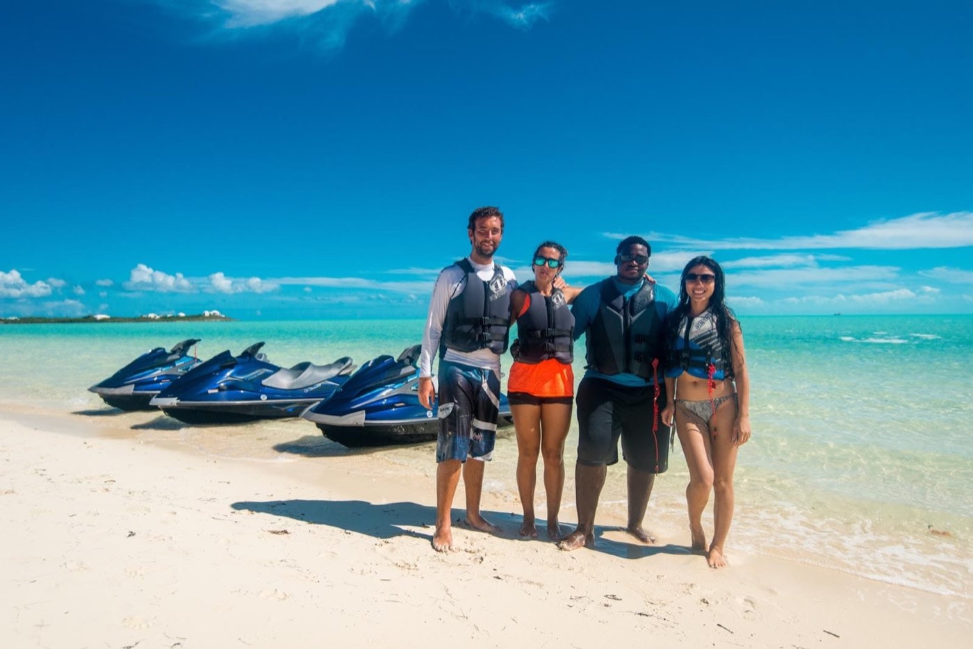 A group of riders standing on a white sand beach beside their jet skis