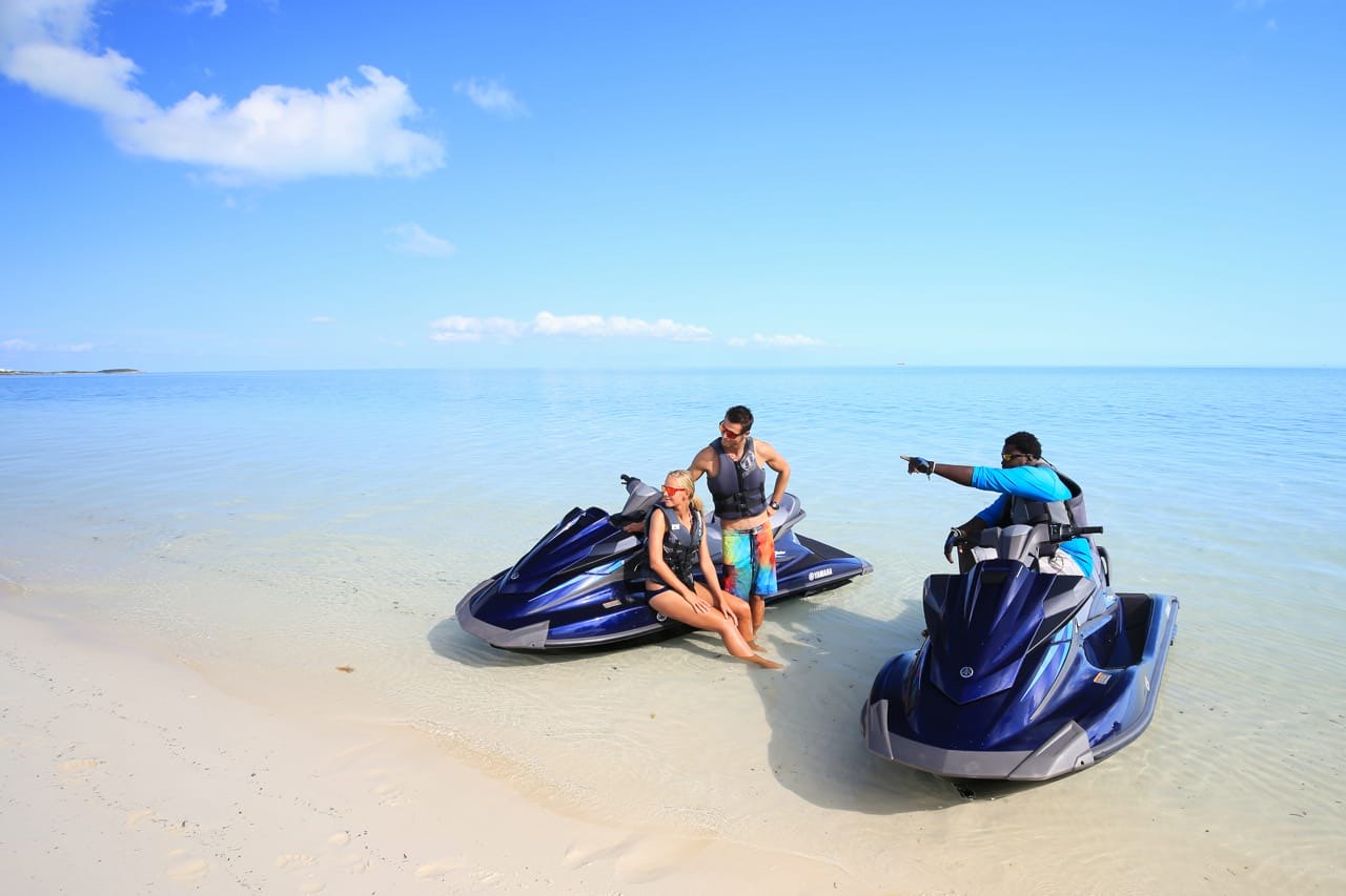 A guide pointing out the route to riders on jet skis pulled up on a white sand beach